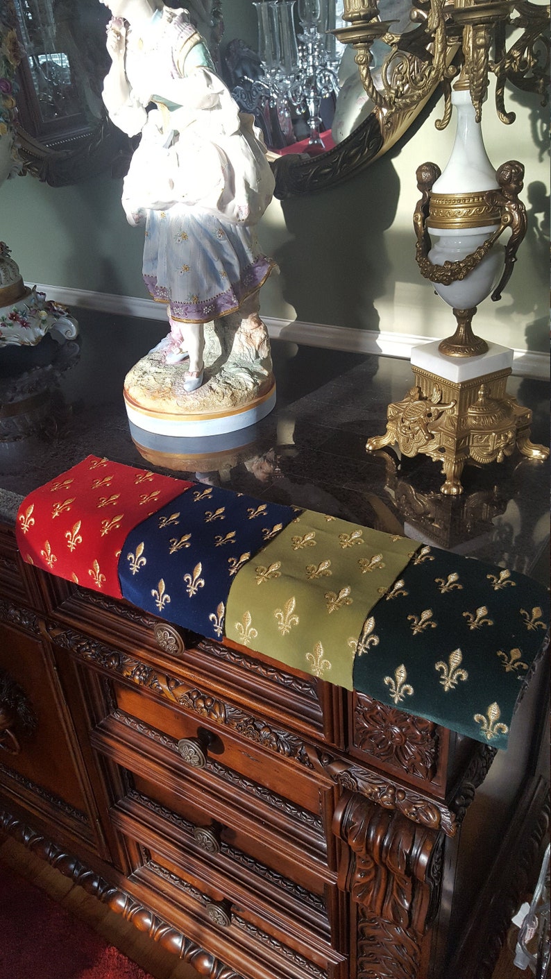 May include: A wooden dresser with four drawers and ornate carvings. A decorative runner with a repeating pattern of gold fleurs-de-lis on red, blue, green, and black velvet fabric is placed on top of the dresser.