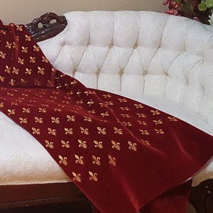 May include: A white tufted chaise longue with a red velvet throw blanket featuring a gold fleur-de-lis pattern.