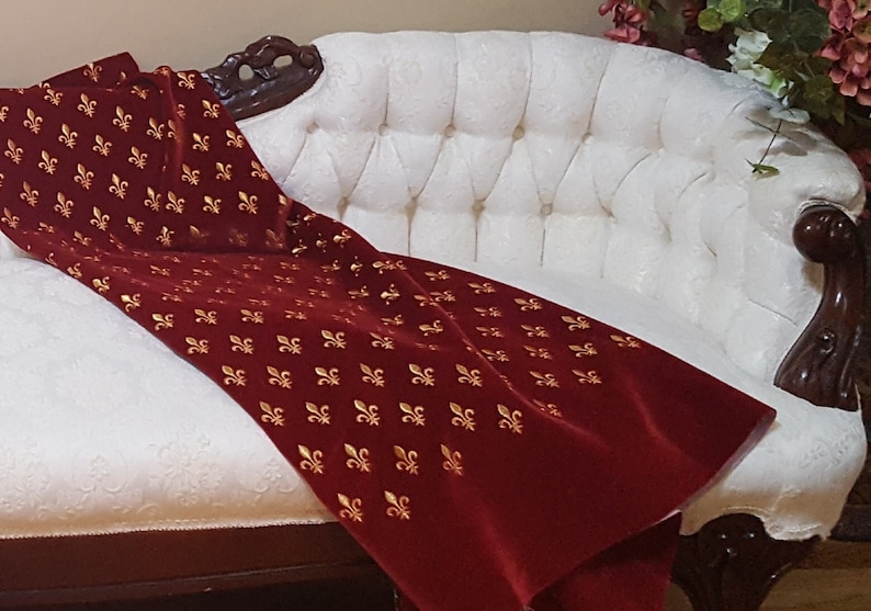 May include: A white tufted chaise longue with a red velvet throw blanket featuring a gold fleur-de-lis pattern.
