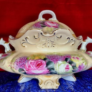 May include: A vintage porcelain serving dish with a lid, featuring a floral design of pink and white roses and purple flowers. The dish has a gold trim and three feet.