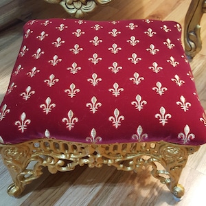 May include: An ornate footstool with a gold-coloured frame and a red velvet cushion featuring a repeating pattern of gold fleur-de-lis.
