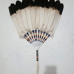May include: A decorative fan featuring white feathers with black tips. The handle is decorated with colourful beads and a tassel. The fan is set against a white textured wall, highlighting its detailed design and craftsmanship.
