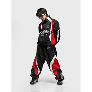 Peut inclure: Ensemble de survêtement noir et rouge avec des détails blancs. La veste présente un logo sur la poitrine et un petit emblème sur la manche. Le pantalon a des panneaux rouges sur les côtés et un logo près de la cheville. Le costume est porté avec des lunettes de soleil.