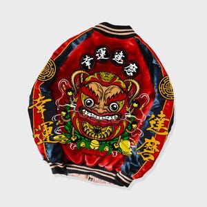 Op de afbeelding: Een rode satijnen bomberjack met een zwart en gouden ontwerp. De jas heeft een groot, rood, cartoonachtig gezicht met een gouden rand en een gouden bel aan elke kant. De jas heeft ook Chinese karakters in goud.