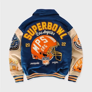 Może przedstawiać: Niebieska i beżowa kurtka bejsbolowa z pomarańczowymi i białymi haftowanymi detalami. Kurtka zawiera napis "SUPERBOWL Los Angeles 20 MPS ORG 22" oraz kask do futbolu z napisem "MPS ORG" z przodu. Rękawy mają napis "Memphis" i "origin" z tyłu.