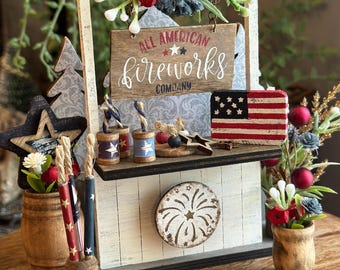 May include: A distressed white wooden display featuring a sign that reads "All American Fireworks Company." The display includes miniature fireworks, an American flag, and decorative floral arrangements. The overall theme is patriotic.