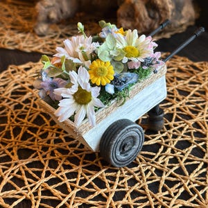 May include: A miniature white wooden wheelbarrow filled with an assortment of colorful artificial flowers. The flowers include white daisies, yellow blooms, and blue and pink accents. The wheelbarrow has a rope trim and black wheels, set on a woven mat.