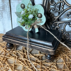 May include: A green shamrock-shaped wooden sign with a floral arrangement and decorative accents. The sign is displayed on a black wooden pedestal with a distressed finish.