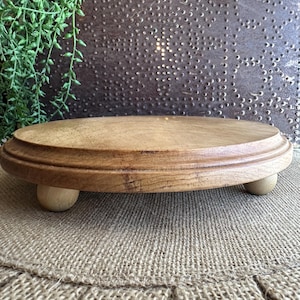 May include: A round, light brown wooden pedestal with a raised edge, supported by three small, rounded feet. The pedestal sits on a burlap surface, with green foliage in the background. The wood has a natural grain.