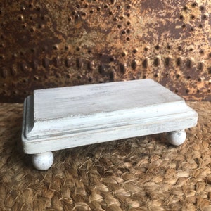 May include: White wooden pedestal with a distressed finish and rounded legs. The pedestal is small and would be perfect for displaying a small item.