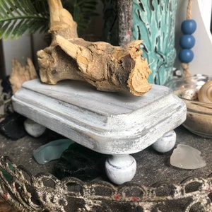 May include: A white wooden pedestal with rounded edges and four white ball feet. A piece of driftwood is placed on top of the pedestal.