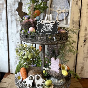 May include: A two-tiered metal tray decorated with a spring and Easter theme. The top tier features a wooden bunny, a basket filled with faux eggs and greenery, and a chalkboard sign that reads "Happy Easter". The bottom tier features a wooden bunny, a nest with faux eggs, and a carrot.