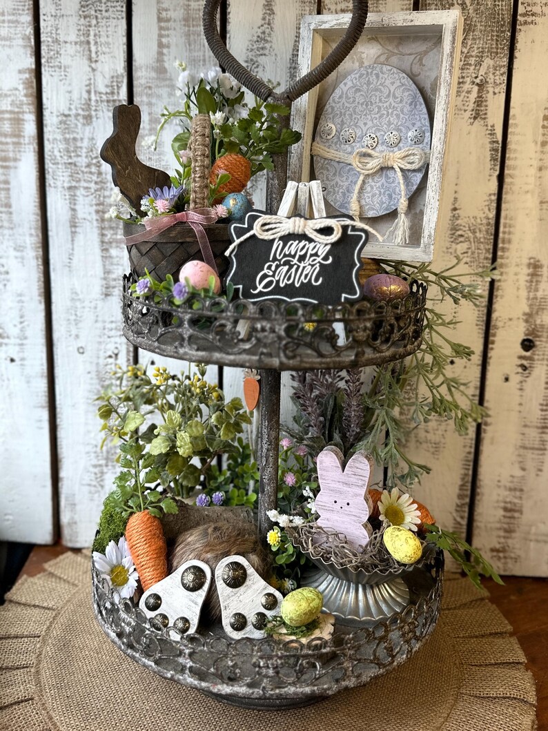 May include: A two-tiered metal tray decorated with a spring and Easter theme. The top tier features a wooden bunny, a basket filled with faux eggs and greenery, and a chalkboard sign that reads "Happy Easter". The bottom tier features a wooden bunny, a nest with faux eggs, and a carrot.