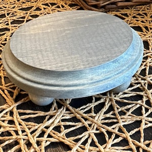 May include: A round, gray-stained wooden riser featuring a smooth, flat top surface with a beveled edge and three small, rounded feet. The wood grain is visible through the gray finish, providing a textured appearance to the circular stand.