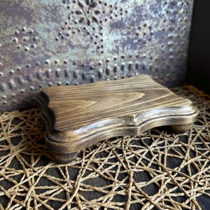 May include: A small, stained wooden riser with a decorative edge and four small feet. The riser is a warm brown color and sits on a woven mat. The background features a textured, metallic surface.