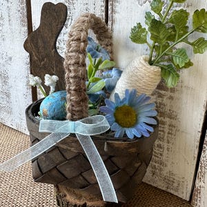 May include: A brown woven basket with a blue ribbon, filled with blue flowers, green leaves, and a white carrot. A wooden bunny cutout is attached to the side of the basket.