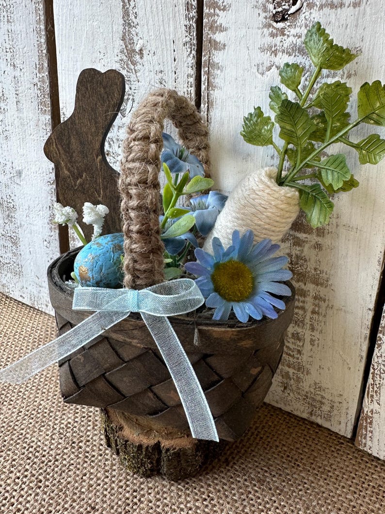 May include: A brown woven basket with a blue ribbon, filled with blue flowers, green leaves, and a white carrot. A wooden bunny cutout is attached to the side of the basket.