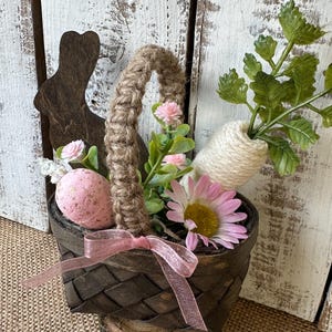 May include: A small brown woven basket with a pink ribbon, filled with pink and white flowers, a pink Easter egg, and a white carrot-shaped twine decoration. The basket is sitting on a wooden stump.