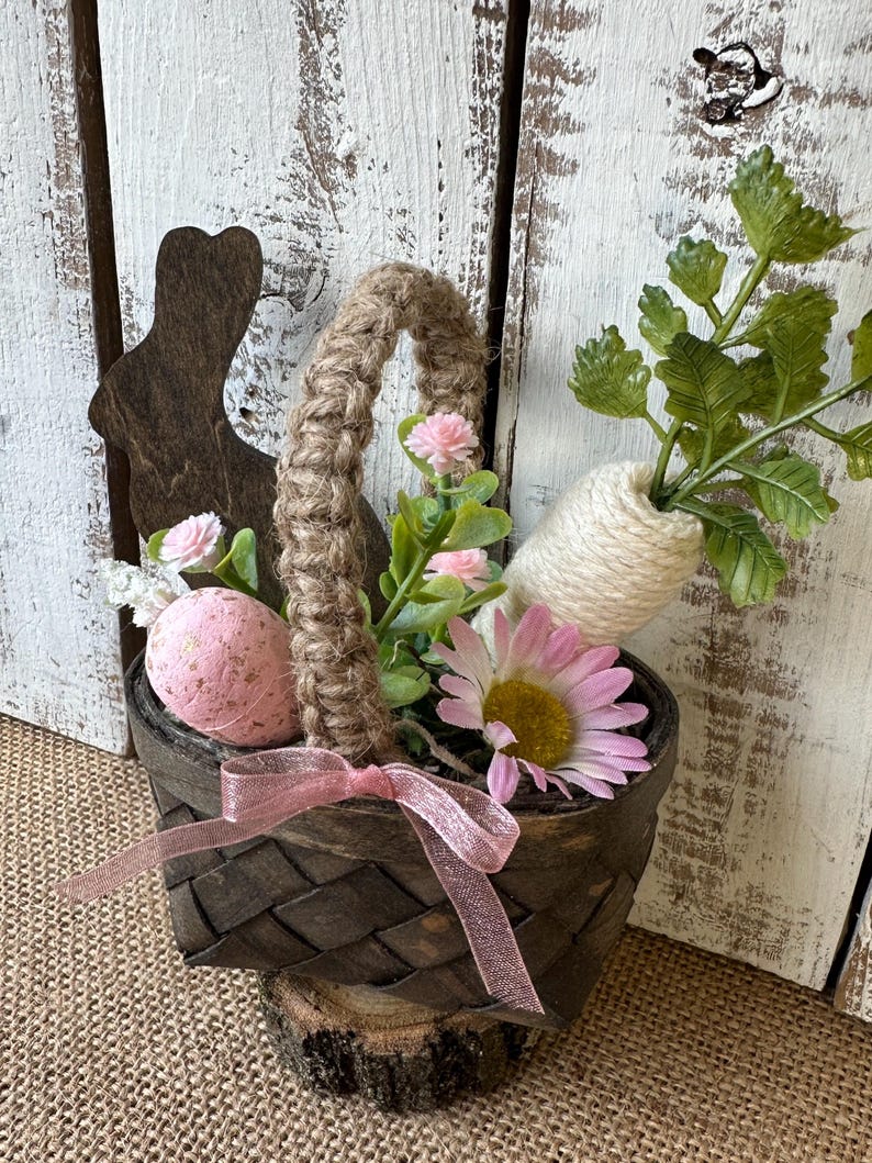 May include: A small brown woven basket with a pink ribbon, filled with pink and white flowers, a pink Easter egg, and a white carrot-shaped twine decoration. The basket is sitting on a wooden stump.