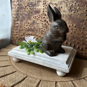 May include: A white wooden tiered stand with a black cast iron bunny figurine on top. The stand has two tiers and is decorated with a whitewash finish. The bunny is sitting on its haunches and has a sweet expression. The stand is sitting on a burlap rug.