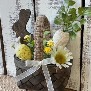 May include: A brown woven basket with a burlap handle, filled with faux flowers, greenery, and yellow Easter eggs. The basket is sitting on a wooden log and has a white ribbon tied around it.
