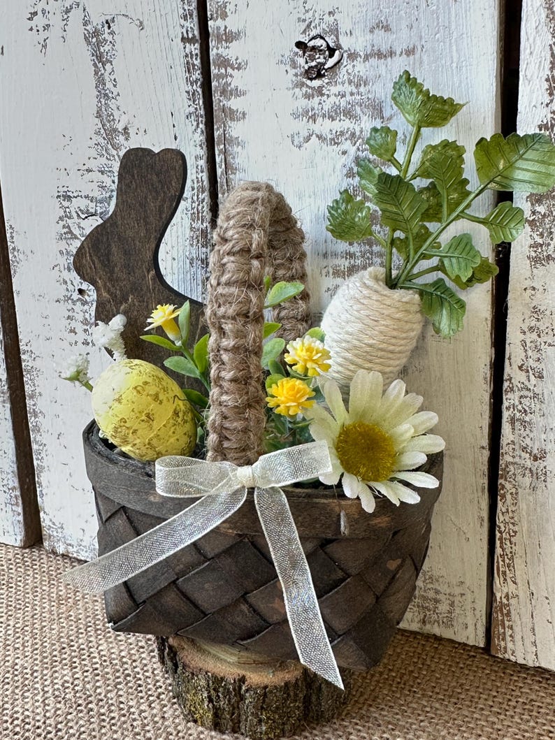 May include: A brown woven basket with a burlap handle, filled with faux flowers, greenery, and yellow Easter eggs. The basket is sitting on a wooden log and has a white ribbon tied around it.