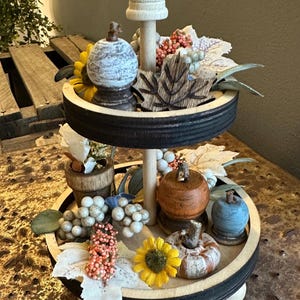 May include: A two-tiered decorative tray with fall-themed accents. The tray features small pumpkins, sunflowers, and leaf-shaped decorations. The tray is made of wood and has a black rim. The overall color scheme includes orange, yellow, and blue.