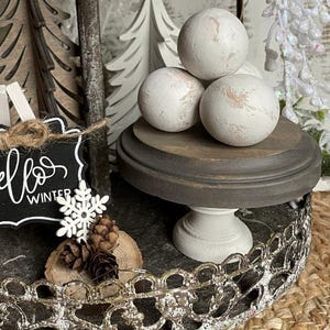 May include: A decorative tiered tray with winter-themed accents. Features white distressed spheres on a wooden pedestal, a chalkboard sign with "hello winter" text, wooden trees, and pinecones. The tray has a decorative metal edge.