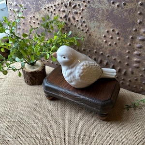May include: A small, white ceramic bird figurine with a brown beak sits on a dark wooden pedestal. The bird is detailed with textured feathers. A small green plant in a wooden pot is to the left, with a textured metal background.