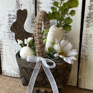 May include: A small brown woven basket with a white ribbon and a white egg inside. The basket is filled with white flowers and green leaves. The basket is sitting on a wooden log.