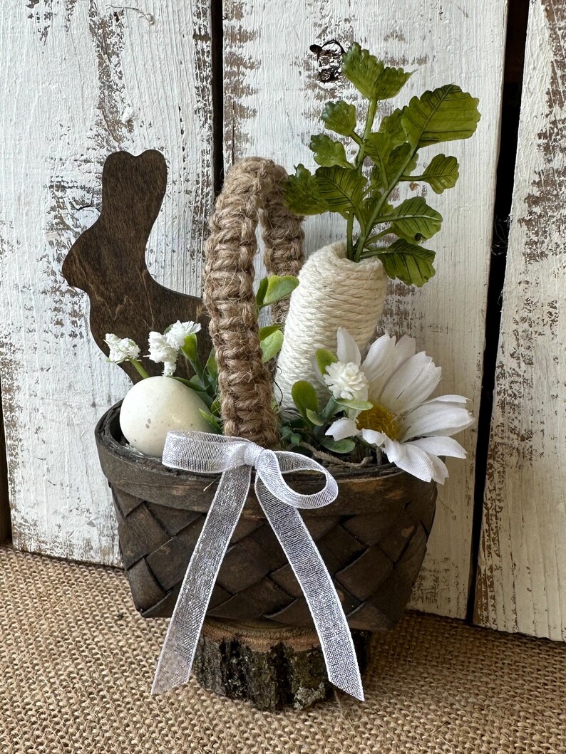 May include: A small brown woven basket with a white ribbon and a white egg inside. The basket is filled with white flowers and green leaves. The basket is sitting on a wooden log.