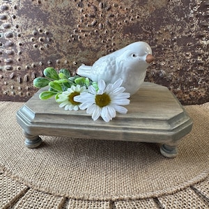 May include: A white ceramic bird figurine sits on a gray wooden tiered stand with four legs. The stand is decorated with a distressed finish and is surrounded by white daisies and green foliage.