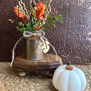 May include: A small, rustic, metal vase with a brown twine bow, filled with a bouquet of orange and brown dried flowers, sits on a wooden stand with a white pumpkin in the foreground.
