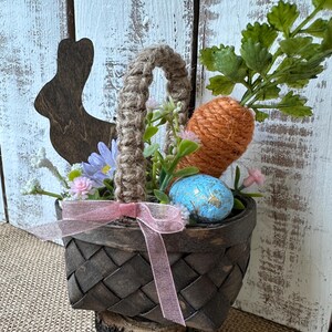 May include: A small brown wicker basket with a burlap handle filled with faux greenery, a carrot, and a blue Easter egg. The basket is sitting on a small wooden log.