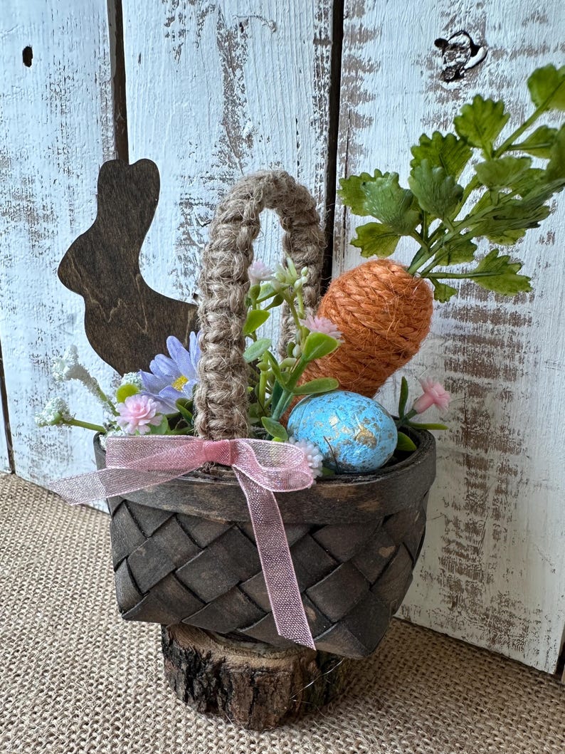 May include: A small brown wicker basket with a burlap handle filled with faux greenery, a carrot, and a blue Easter egg. The basket is sitting on a small wooden log.