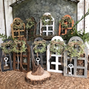 May include: Set of eight wooden window ornaments with a rustic farmhouse style. Each window features a key, a wreath, and a distressed paint finish. The windows are arranged on a woven rug with a wooden slice in the center.