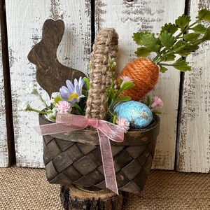 May include: A small brown wicker basket with a pink ribbon, filled with a carrot, a blue speckled egg, and a wooden bunny cutout. The basket is sitting on a wooden log and surrounded by green leaves and pink flowers.