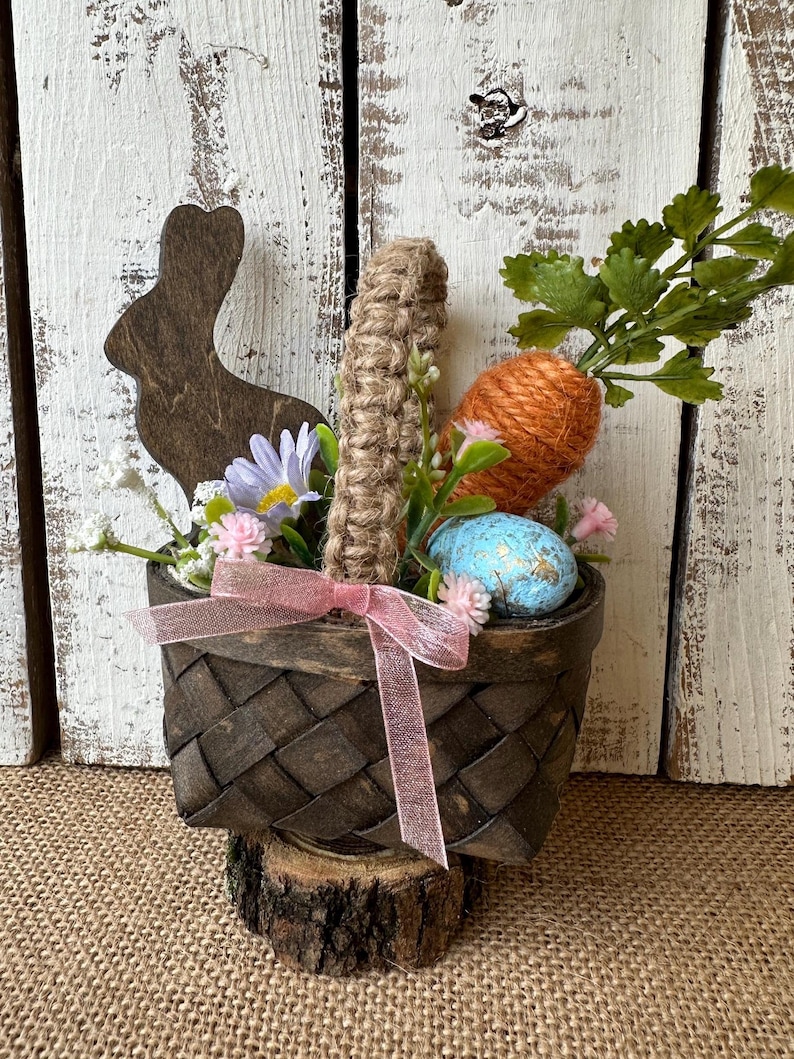 May include: A small brown wicker basket with a pink ribbon, filled with a carrot, a blue speckled egg, and a wooden bunny cutout. The basket is sitting on a wooden log and surrounded by green leaves and pink flowers.
