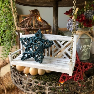 May include: A white miniature swing set with a blue star-shaped cushion and a red star-shaped cushion. The swing set is hanging from a metal stand with a rustic finish. The stand has a top tier with a green plant and a metal birdcage with lights. The bottom tier has a red and white floral arrangement in a metal container with the text "1776" on the side.
