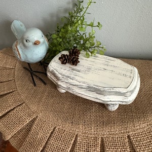 May include: A small white wooden pedestal with four legs, distressed with a white paint finish. The pedestal is sitting on a burlap rug with a blue bird figurine and a small sprig of green foliage.