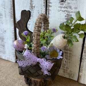 May include: A small wooden basket with a burlap handle, filled with faux flowers, a carrot, and a painted Easter egg. A wooden bunny cutout is attached to the side of the basket.