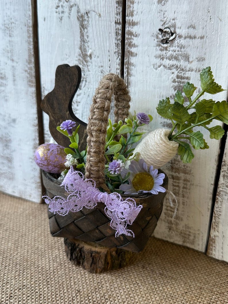 May include: A small wooden basket with a burlap handle, filled with faux flowers, a carrot, and a painted Easter egg. A wooden bunny cutout is attached to the side of the basket.