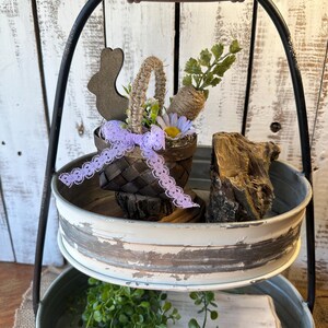 May include: A two-tiered metal serving tray with a distressed white finish. The top tier features a small woven basket with a purple ribbon, a wooden bunny, and a faux carrot and greenery. The bottom tier has a wooden board and green foliage.