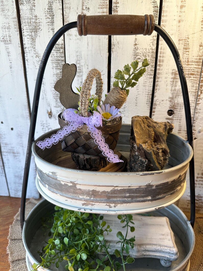 May include: A two-tiered metal serving tray with a distressed white finish. The top tier features a small woven basket with a purple ribbon, a wooden bunny, and a faux carrot and greenery. The bottom tier has a wooden board and green foliage.