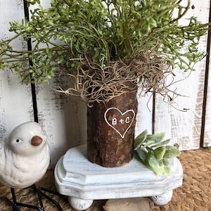 May include: A white wooden pedestal with a log vase with green leafy branches and the initials "B+C" carved into the side. The vase is sitting on a white wooden pedestal with three legs.