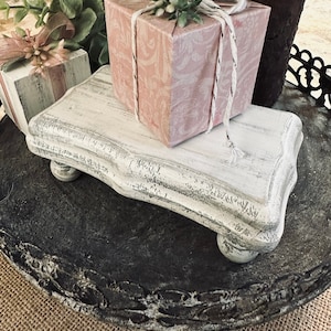 May include: A white wooden tiered stand with a distressed finish. The stand has a scalloped edge and four turned legs. It is decorated with two pink gift boxes, one with a white bow and the other with a pink bow.