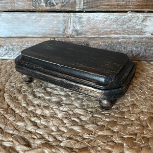 May include: A small black wooden pedestal with a rectangular top and rounded legs. The pedestal is distressed and has a rustic finish.