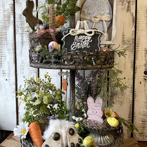 May include: A two-tiered metal serving tray decorated with a rustic, distressed finish. The top tier features a wooden bunny, a basket filled with Easter eggs, and a sign that reads "Happy Easter". The bottom tier has a white bunny, a nest with eggs, and greenery.