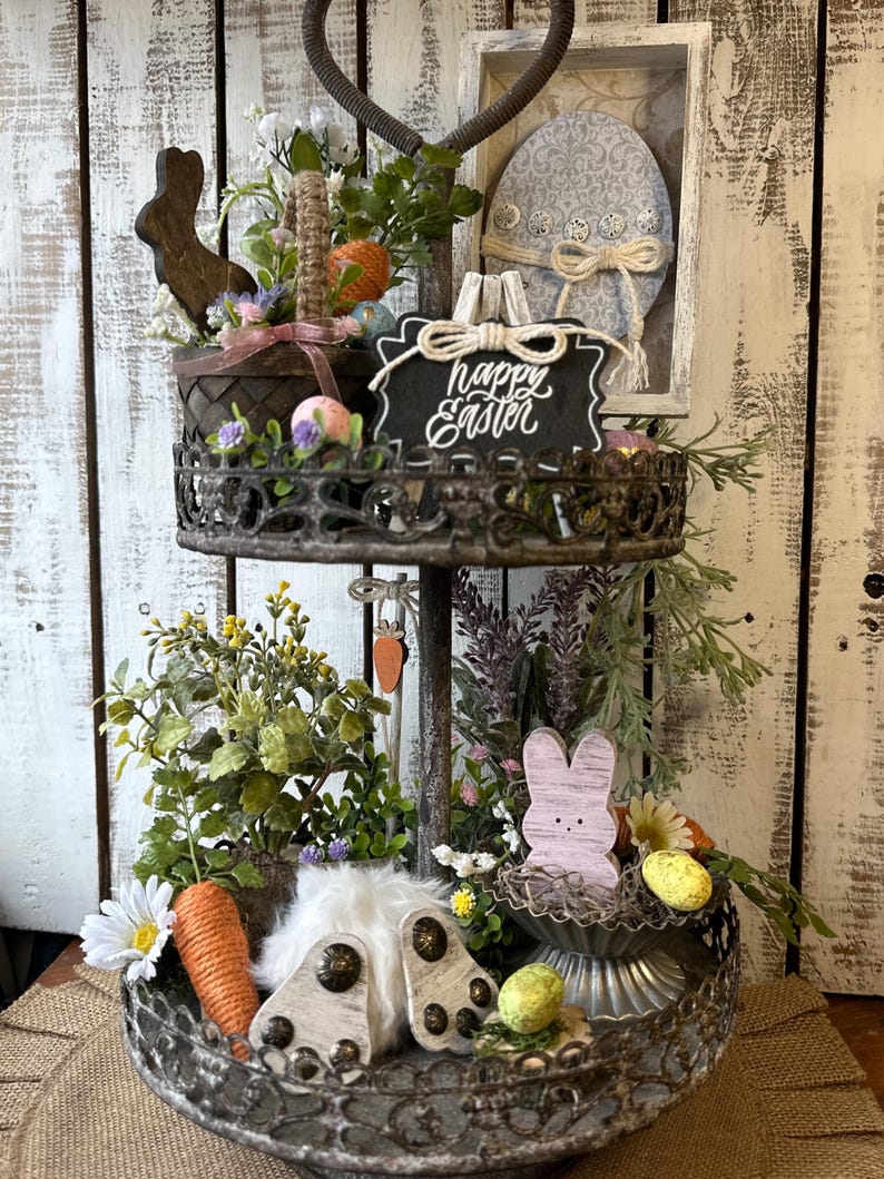 May include: A two-tiered metal serving tray decorated with a rustic, distressed finish. The top tier features a wooden bunny, a basket filled with Easter eggs, and a sign that reads "Happy Easter". The bottom tier has a white bunny, a nest with eggs, and greenery.