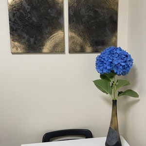 May include: Two abstract paintings with black and gold textured paint on canvas, hanging on a white wall. A blue hydrangea flower in a black and gold vase sits on a white table with a black chair.